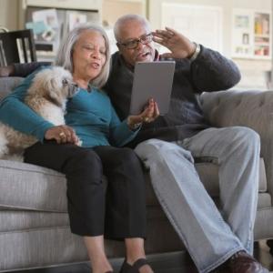 Two people and a small white dog sitting on a couch, looking at a tablet device.