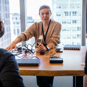 A person demonstrating a device to others, a large open window behind them.