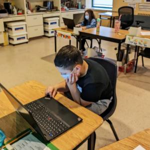 child in a mask and a black t-shirt sits at a desk with a lenovo laptop