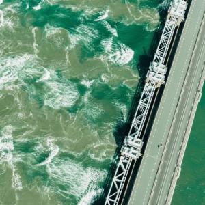 Aerial view of a large dam with green water on one side and cloudy green turbulent water on the other.