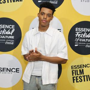 A person standing, facing the camera in front of a yellow backdrop with Essence Film Festival logos.