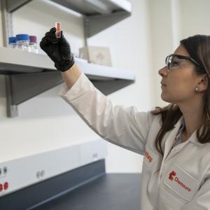 A person wearing protective lab coat, glasses and gloves, inspecting a fluid in a small tube in a laboratory setting.