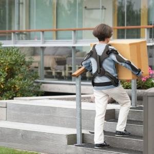a petite person wearing an 'exoskeleton' carrying a box up steps