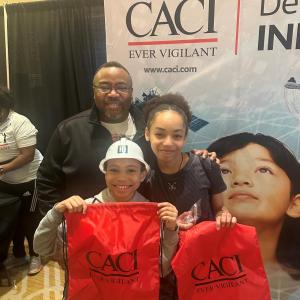 A group of three pose in front of a CACI banner, holding a CACI bag.