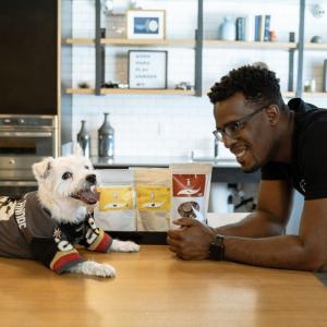 a small white dog in a sports jersey, sitting on a table with Eric Adams, in a black t-shirt, looking on