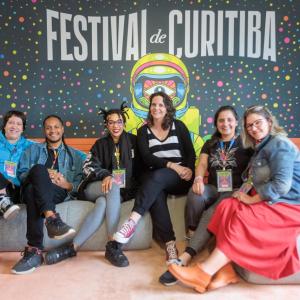 People seated on cushions, a large "Festival de Curitiba" sign behind them.