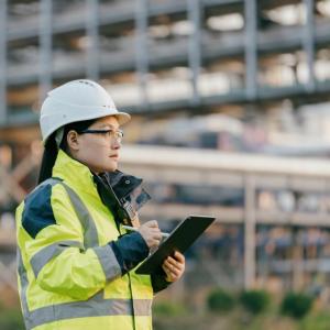 A person wearing high-vis clothing and hard hat, using a tablet. Large scaffolding behind them.