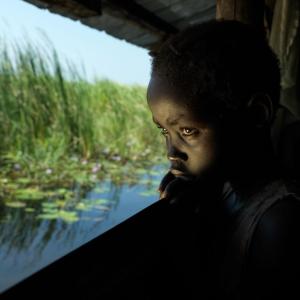 Ten-year-old, Nyma waits for her mother to return home from serving tea to the local authority. They both live in an abandoned classroom in the school she used to attend before it closed due to the floods. Using locally sourced grass, they built a floating pontoon on top of the concrete classroom floor to mitigate the rising flood water. Nyma gets anxious when her mother is away and misses her old home.