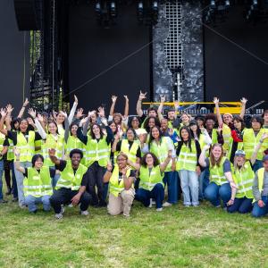 Large group wearing bright yellow safety vests poses together with raised hands in front of an outdoor concert stage.