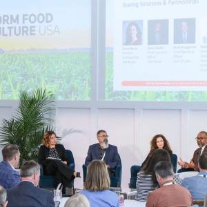 Panel of five seated at the front of a room of others listening to one speak.