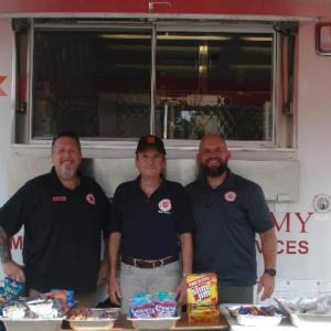 Three people standing in front of a box truck with a table of food in front.