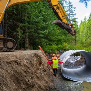 A large machine lifting a metal culvert as workers in a ditch position it.