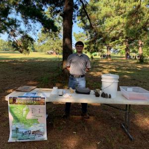 A person standing in a park behind a table with presentation materials and a map of Louisiana.