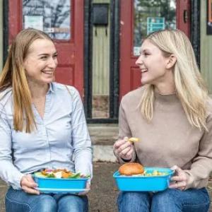 co-founders Jacquie Hutchings and Kayli Dale sitting on a front porch, eating out of blue containers