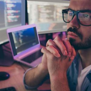 A person with folded hands looking at multiple monitors on a desk.