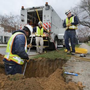 Utility workers watch as on measures in a dirt hole next to a sidewalk.