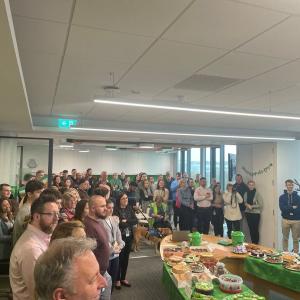 A room packed with people standing, an oval table with food on it. 