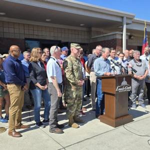 A group meeting behind a person speaking at a podium outside.