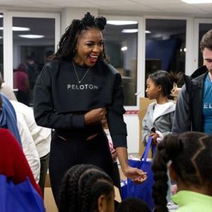 Two people wearing Peloton shirts. A small group of children in front of and behind them each carrying a blue bag.
