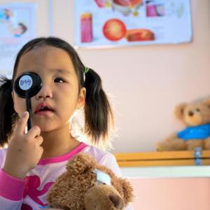 A child holding a tool to cover one eye. Holding a teddy bear with the other hand.