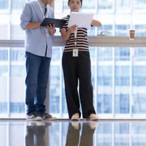 Two people standing in an open room with tall windows, looking at the same paper.