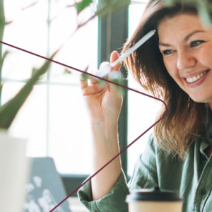 A smiling person looking at a laptop, a pen by their head.