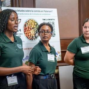 Three people in green shirts presenting. A display board behind them.