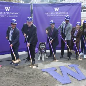 seven people in hard hats holding shovels dug in to sand around a large "W" and one very good dog in the middle
