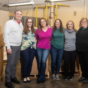Six people posed with arms around each other. Large spools of thread to the side and an Eastman Box on the other side.