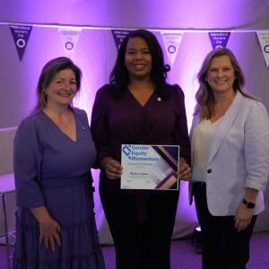 Three people standing together with a purple background, one holding a certificate