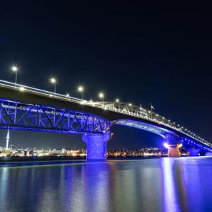 night panorama of Auckland Harbour Bridge over an expanse of water and is lit up in blue lights