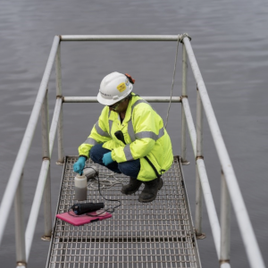 worker in safety gear crouched at the end of a metal catwalk over a body of water
