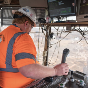 worker in a bright orange shirt and a hard hat at the controls of a large vehicle
