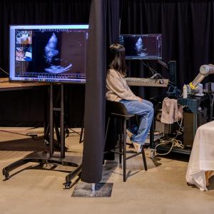 A person on a table with another sitting nearby. A medical imaging device on their chest. Another person behind a curtain viewing the images.