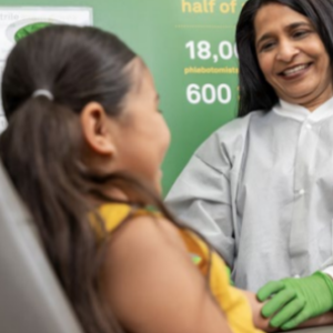 A smiling adult in protective gear looking at a child, seated.