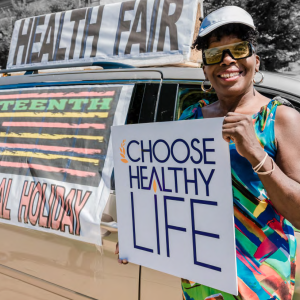a person holds a sign "choose healthy life" infront of a vehicle with "health fair" on the roof and a sign over the window "...teenth...Holiday"