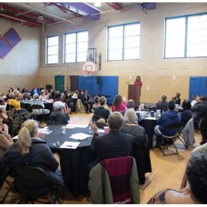 A room of seated people in a gymnasium. A person speaking at a podium at the front.
