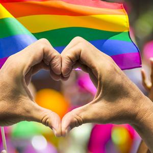 fingers forming a heart with rainbow flags in the background