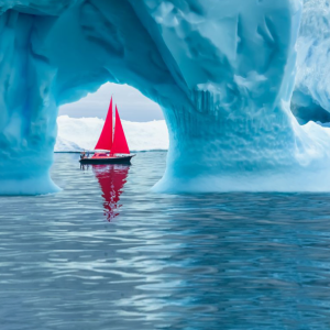 A boat with red sails framed in a small opening in an iceberg on a body of water.