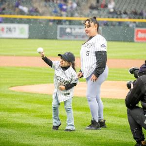 A child throwing a pitch on a baseball field, with an adult behind them and a photographer shown