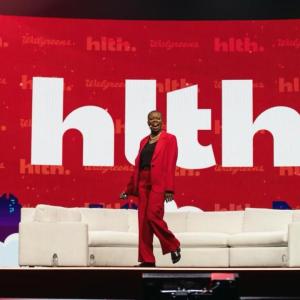 Tracey Brown on a large stage with a white couch and large digital display behind.