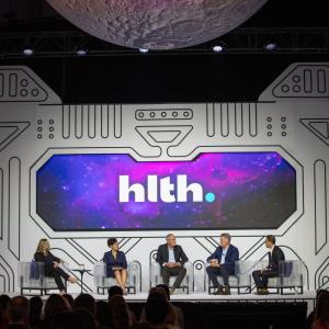 A panel of five on a stage, a digital display behind.