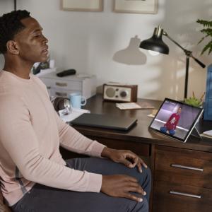 a person sitting at a home office desk, eyes closed, a meditation program playing on the device on their desk