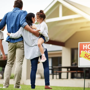 A family standing in front of a hours with a "SOLD" sign in front of it