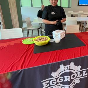 A person standing with food behind a table with a cloth that says "The Eggroll Factory"