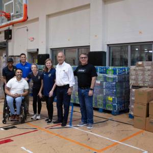 People posed in front of pallets of boxes of food, water and supplies in a gymnasium.