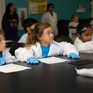 Three students in lab coats and goggles sitting in a science lab.