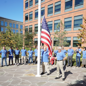 Veterans Day 2021 flag-raising ceremony at Illumina headquarters. Two males are raising the flag and a group of Illumina veterans are in a semi-circle around the back of the flag pole. The crowd is made up of 12 men and women.