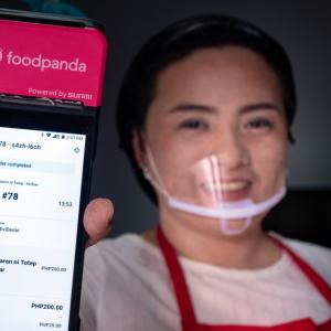 woman in red apron and a plastic face mask holds up a phone featuring the food panda app