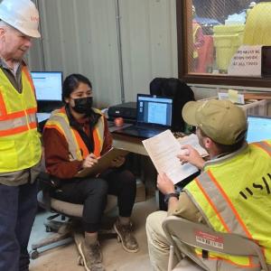 Three workers in an office wearing safety gear, one holds a clipboard and the other a stack of papers